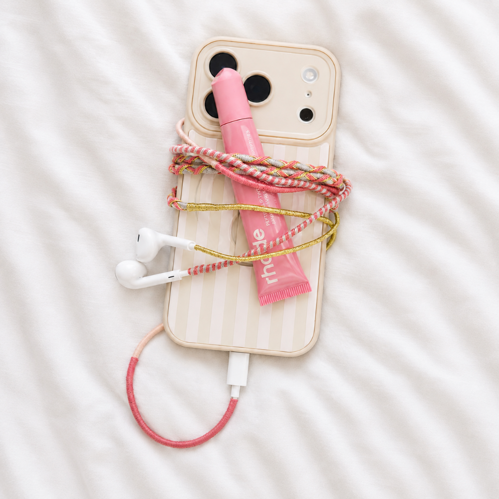 A styled flat-lay of a cream-colored striped phone case with a pink and white braided cable, a pink lip balm tube, and white wired earbuds on a white textured fabric background.