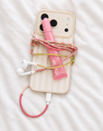 A styled flat-lay of a cream-colored striped phone case with a pink and white braided cable, a pink lip balm tube, and white wired earbuds on a white textured fabric background.